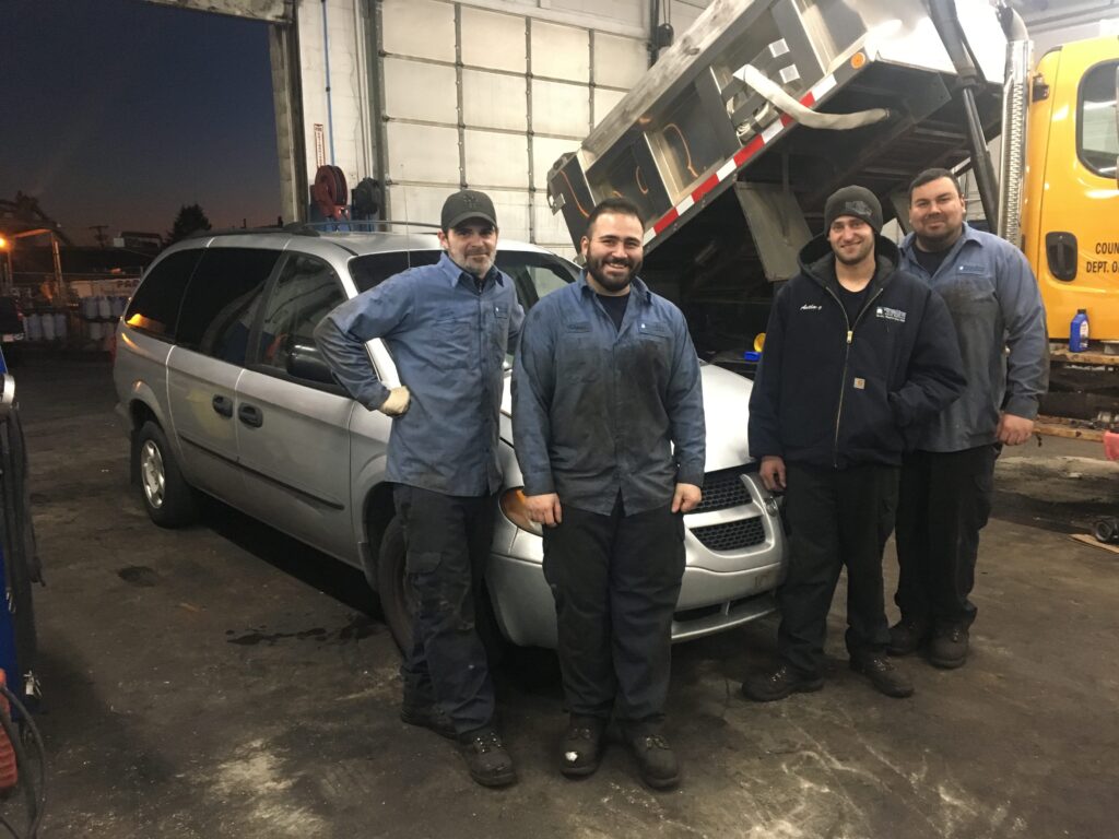 A picture of people smiling at a mechanic facility with trucks and vans in the background